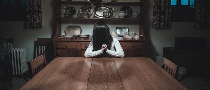  A woman with hair completely covering her face sitting alone at the dining room table in a dimly lit room. You get the sense that this is super creepy and that you don't want to stay long. 