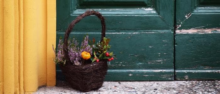  A brown wooden flower basket with an assortment of flowers sits in front of a door. The door is green and the walls surrounding it are yellow. The paint is peeling. 